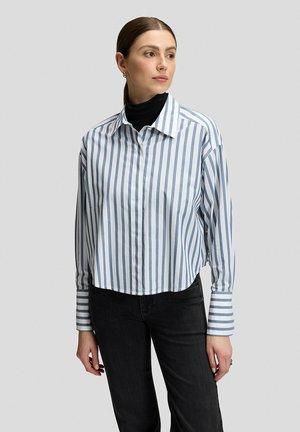 Donna in piedi rivolta a sinistra, indossa una camicia a righe verticali blu e bianche sopra un dolcevita nero e dei jeans neri su uno sfondo neutro.