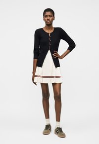 Femme portant un cardigan noir boutonné, une jupe plissée blanche avec des rayures rouges, des chaussettes blanches et des baskets vert olive, debout, main sur la hanche.