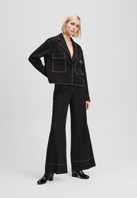Black cropped blazer with white stitching, dual front pockets, paired with black wide-leg trousers and glossy black ankle boots.