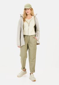 Lichtgrijze gewatteerde jas, crèmekleurige top, olijfgroene cargopantalon en lichte sneakers. Inclusief een groene pet met een cirkelvormig logo. Casual outfit.