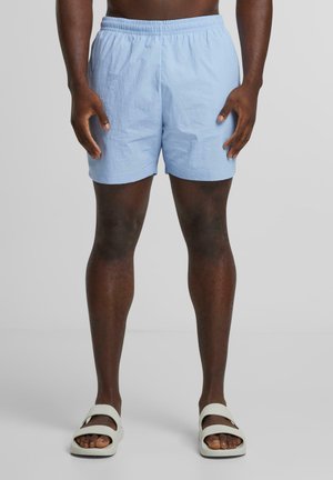 Shorts bleu clair, arrivant à la taille, fabriqués dans un tissu léger avec une taille élastique. Portés avec des sandales pâles. Texture lisse, design décontracté.