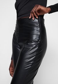 Svarta byxor i konstläder med hög midja, vertikala sömdetaljer och en sidogångjärn. Slät textur och skräddarsydd passform.