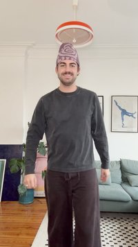 Mørkegrå langærmet sweatshirt og sorte bukser med vide ben. Den lilla hat har et snurrende mønster. Indendørs miljø med grøn plante og møbler.