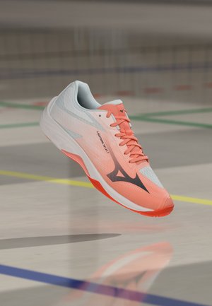 Könnyű korallszínű és fehér sportcipő fekete logóval, amely a beltéri pálya színes határvonalai felett lebeg.