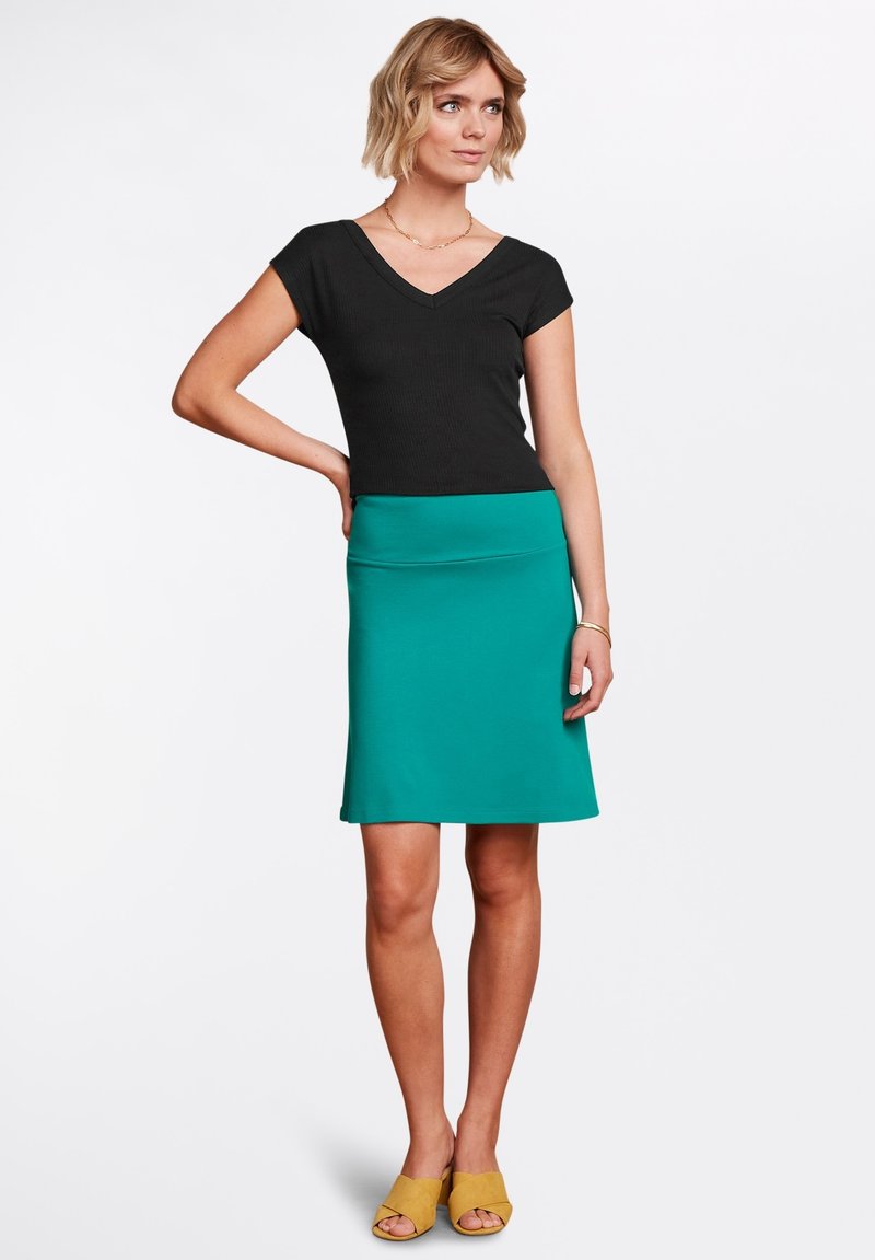 Sort ribbet V-hals top og teal, knælang nederdel. Modellen har gule åbne sko på. Glat stof med tætsiddende design.
