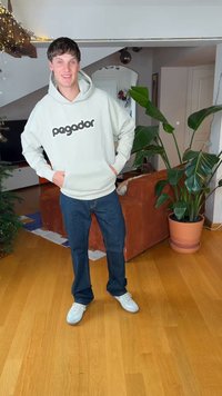 Hellgrauer Hoodie mit "pagador" in Schwarz, dunkelblaue Jeans und weiße Turnschuhe. Steht auf Holzboden in der Nähe von grünen Pflanzen.