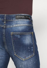 Siniset farkut, joissa on kulunut pinta, kontrastiväriset keltaiset tikkaukset ja koristeellisia maalikosketuksia. Tuote on merkitty "Alessandro Zavetti."