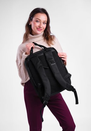 Černý batoh s polstrovanými popruhy a strukturovaným designem, který má prošívaný přední díl a odolné materiály. Držen jedincem v svetru.