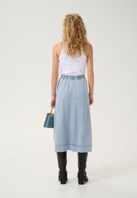 Nainen, jolla on kihara vaaleanblondi hiukset, pukeutuneena valkoiseen toppiin, vaaleansiniseen farkku-midi-hameeseen ja mustiin polvipitkiin saappaisiin, pitäen kädessään pientä sinistä käsilaukkua.