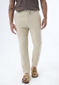 Beige Leinenhosen mit glatter Textur, taillierter Passform, verstellbarem Bund und Fronttaschen, kombiniert mit sandfarbenen Riemensandalen.