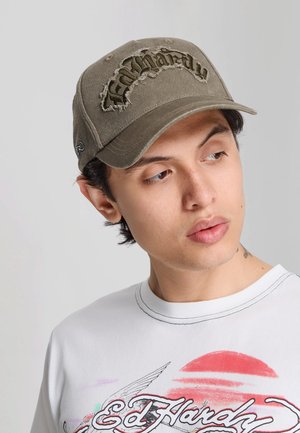 Młody mężczyzna noszący znosioną zieloną czapkę Ed Hardy oraz białą koszulkę Ed Hardy z kolorowym nadrukiem graficznym, patrzy w lewo.