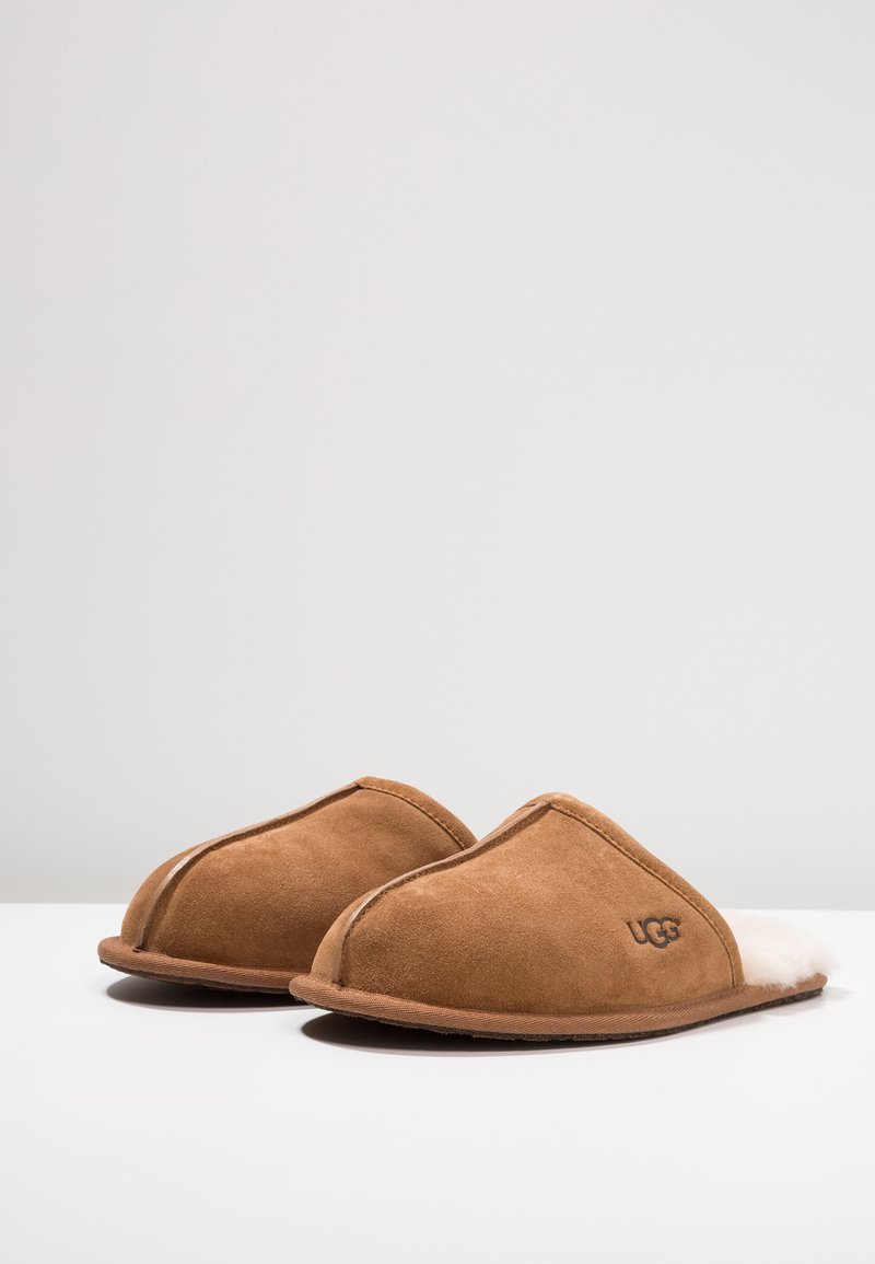 Braune Wildleder-Schlappen mit einem weichen weißen Fellfutter, geschwungenem Design und einer flachen Gummisohle. UGG-Logo auf der Seite geprägt.