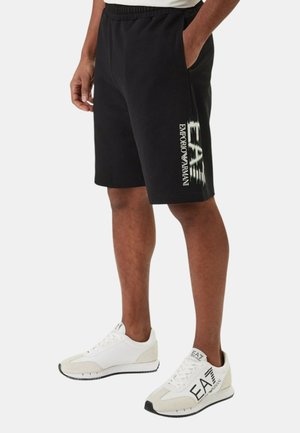 Uomo che indossa pantaloncini neri EA7 Emporio Armani e sneakers bianche con marchio EA7, in piedi contro uno sfondo chiaro e semplice.