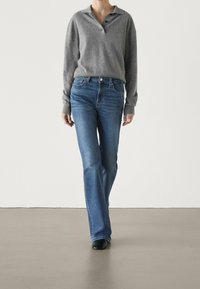 Grå långärmad polo skjorta med krage och tre knappar, i kombination med blå utsvängda jeans. Tyget ser mjukt och bekvämt ut.