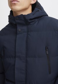 Marineblå pufferjakke med glidelåslomme, trykknapper, justerbar hette og teksturert stoff. Har horisontal søm for isolasjon.