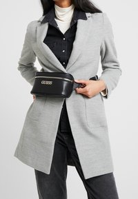 Černá kožená ledvinka s texturovaným povrchem, se stříbrným zipem a logem "GUESS". Nosená přes šedý kabát a černé tričko.