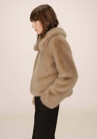 Giacca in pelliccia sintetica beige con colletto tondo, maniche ampie e silhouette corta, indossata sopra un pantalone nero aderente.