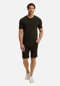 Tmavě zelené tričko s krátkým rukávem a odpovídající šortky z měkkého, hladkého materiálu. Model má na sobě bílé tenisky, ruce v kapsách.