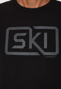 Mustat lyhyet hihapaita, joka on valmistettu puuvillasta ja jossa on suuri harmaa "SKI"-logo ja "J. Lindeberg" -teksti etupuolella. Klassinen pyöreä kaula-aukko.