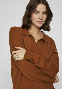 Suéter de punto marrón con cuello, que presenta puños y dobladillo acanalados. La tela parece suave y tiene un corte holgado.