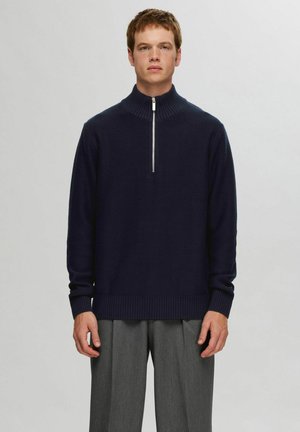 Navy zip-up sweater with high collar, ribbed cuffs and hem, made of soft knit material. Worn with tailored gray trousers.