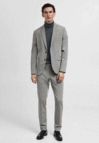 Costume gris clair à coupe ajustée. Comprend un blazer à un bouton et un pantalon assorti, associé à un col roulé gris foncé. Chaussures noires.