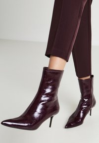 Stivali alla caviglia a punta di un elegante bordeaux scuro, con tacchi alti e sottili, indossati con pantaloni scuri e cropped su un pavimento chiaro.