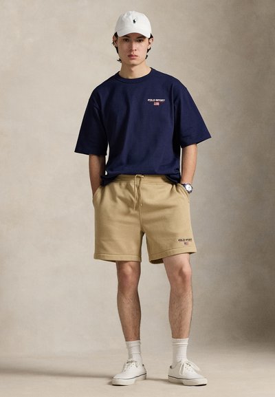 T-shirt bleu marine à manches courtes avec logo "Polo Sport", associé à un short beige à taille élastique. Le modèle porte des baskets blanches et une casquette blanche.