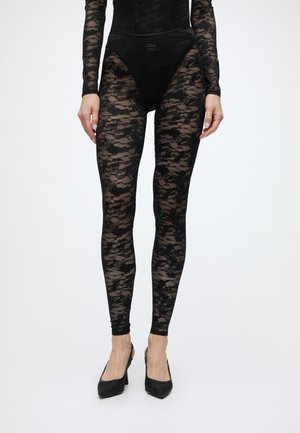 Donna che indossa leggings in pizzo nero trasparente, top abbinato a maniche lunghe, biancheria intima nera e décolleté nere a punta, in piedi eretta.