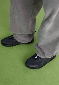 Baskets noires en tissu texturé avec des semelles en caoutchouc, associées à un pantalon en velours côtelé gris, sur une surface verte.