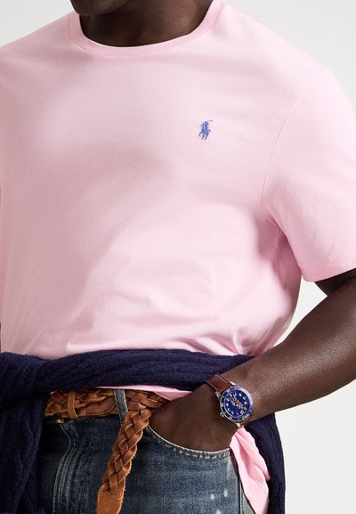 Šviesiai rožinė marškinėlių su apvalia iškirpte, su siuvinėta mėlyna logotipu, derinama su tamsiais džinsais ir austu rudos odos diržu. Mėlynas laikrodis ant riešo.