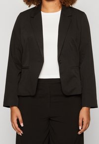 Blazer negro con un diseño entallado, cierre de un solo botón y dos bolsillos frontales. Fabricado en tela suave, combinado con una blusa blanca lisa.