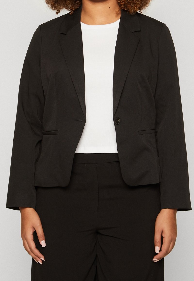 Blazer negro con un diseño entallado, cierre de un solo botón y dos bolsillos frontales. Fabricado en tela suave, combinado con una blusa blanca lisa.