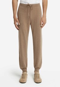 Man med ljusbruna dragstringstrumpor och matchande beige mockaskor, stående mot en vit bakgrund.