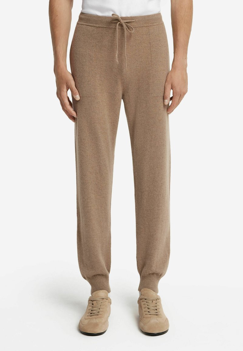 Man med ljusbruna dragstringstrumpor och matchande beige mockaskor, stående mot en vit bakgrund.