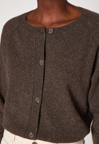 Dunkelbrauner, gemusterter Button-up-Kardigan über hellbeigen Hosen, der den Ausschnitt und den Oberkörper einer Person mit dunkler Haut zeigt.