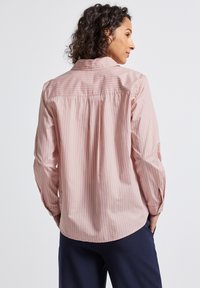 Donna con capelli ricci che indossa una camicia a maniche lunghe a righe verticali rosa chiaro e pantaloni scuri, girata di spalle con le mani in tasca.