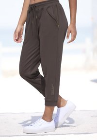 Braune Jogginghosen mit einem Kordelzugbund, seitlichen Reißverschlusstaschen und einem umgeschlagenen Fußdesign. Kombiniert mit weißen Sneakers für einen lässigen Look.
