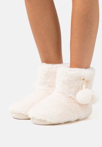 Soft, pink fuzzy slippers with a plush texture, ankle height, rounded toe, and a ribbon-accented pom-pom detail at the side.