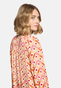 Femme aux cheveux blonds ondulés mi-longs, portant un chemisier à motifs géométriques rouges, oranges et beiges, regardant par-dessus son épaule.