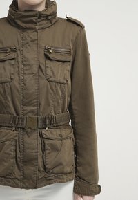Giacca verde oliva realizzata in tessuto con finitura opaca. Presenta un colletto alto, due tasche sul petto con zip, una cintura e epaulettes sulle spalle.