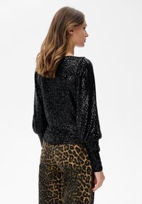 Vrouw met golvend haar draagt een zwart, glinsterend shirt met lange mouwen en een broek met luipaardprint, staande in profiel tegen een effen witte achtergrond.