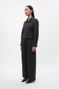 Femeie în picioare purtând o jachetă gri închis cu fermoar și pantaloni largi asortați, cu pantofi negri cu vârf ascuțit, pe fundal alb simplu.