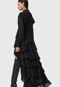 Schwarzes Kleid mit Rüschen und langen Ärmeln, mit mehrlagigen Stufen und einem taillierten Oberteil. Der Stoff ist weich und fließend und betont die Bewegung.