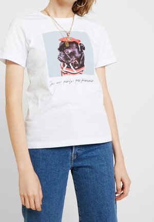 Kvinde iført en hvid t-shirt med et print af en sort mops i en rød baret og en stribet skjorte, med fransk tekst under hunden.