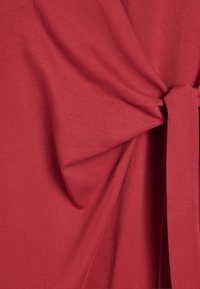 Czerwony materiał o gładkiej fakturze, z wiązanym węzłem po jednej stronie, delikatnie układający się z subtelnymi fałdami.