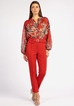 Blouse rouge à motif floral avec un décolleté en V, associée à un pantalon rouge ajusté avec une ceinture et une boucle. La tenue est complétée par des talons beiges.