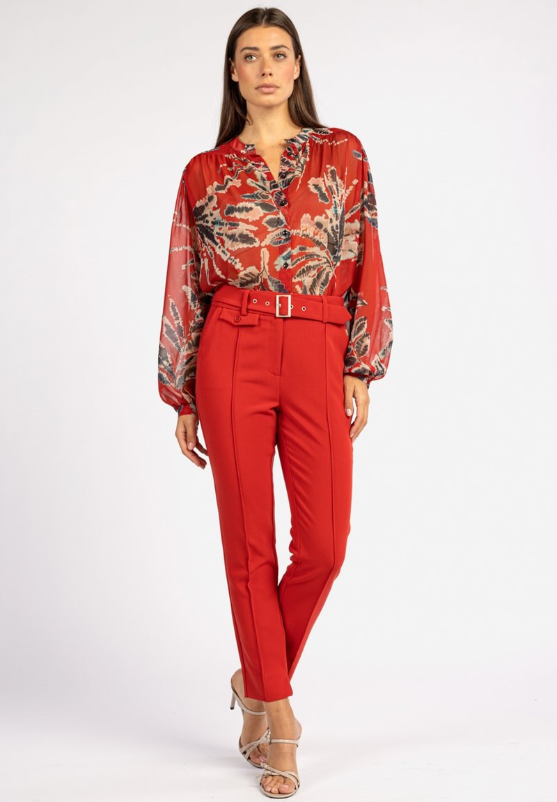 Blouse rouge à motif floral avec un décolleté en V, associée à un pantalon rouge ajusté avec une ceinture et une boucle. La tenue est complétée par des talons beiges.
