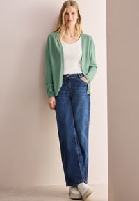 Un cardigan vert clair sur une chemise blanche, associé à un jean large bleu et des baskets blanches. Des textures douces et une coupe décontractée sont évidentes.