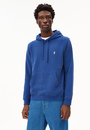 Blauwe capuchontrui van zacht materiaal, met een voorzak en een klein logo op de borst, gecombineerd met blauwe jeans.